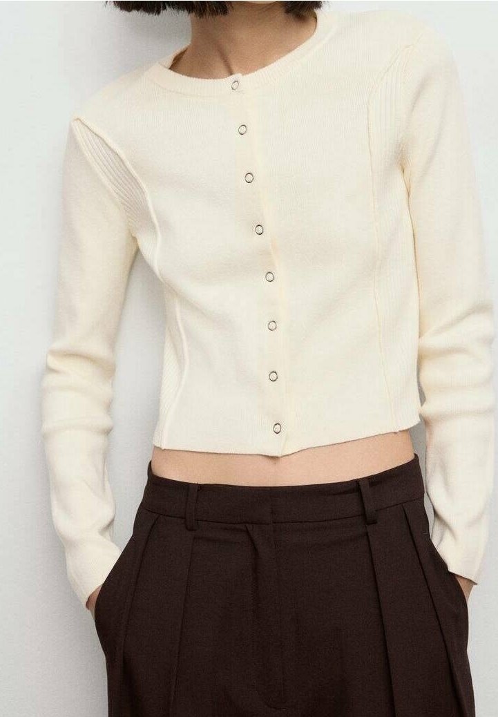 Femme portant un cardigan court couleur crème avec de petits boutons et un pantalon plissé marron foncé taille haute, les mains dans les poches.