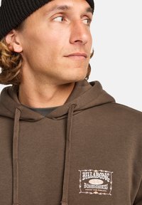 Sweat à capuche marron avec un patch logo. Il comprend une capuche avec cordon de serrage et des poignets côtelés. Le modèle porte un bonnet noir. Fond neutre.