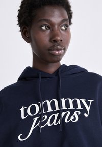 Felpa blu navy realizzata in morbido tessuto, con un logo bianco che recita "tommy jeans" in un carattere corsivo audace. Con corde di regolazione.