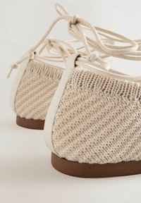 Des chaussures en tissu tissé beige avec un dessus texturé, des accents crème et une semelle marron, ornées de lacets décoratifs noués à l'arrière.