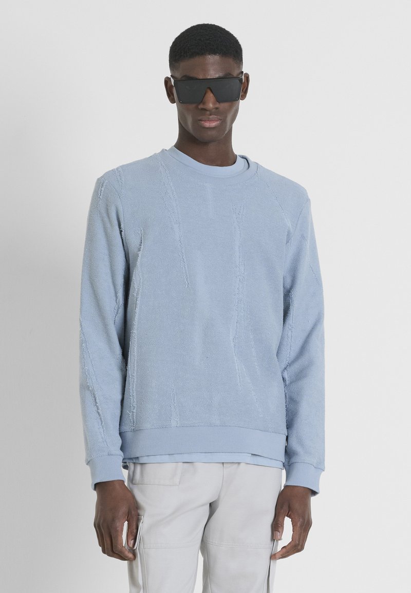Sudadera de color azul claro con un diseño texturizado y desgastado, y puños acanalados. Lleva una camiseta gris claro debajo y está combinada con pantalones claros. Se usan gafas de sol negras.