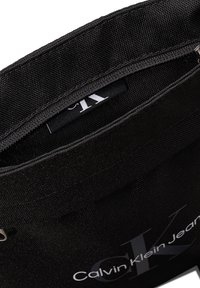 Sort stofpose med lynlås, prydet med et fremtrædende "CK" logo i grå og et label med teksten "Calvin Klein Jeans" indeni.