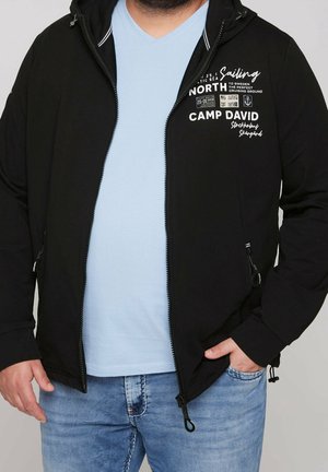 Mann trägt eine offene schwarze Kapuzenjacke mit weißem segelthemenbezogenem Schriftzug über einem hellblauen Hemd und blauen Jeans, eine Hand in der Tasche.