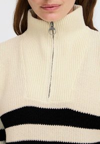 Maglione a maglia color crema con strisce orizzontali nere, colletto alto e chiusura con zip a metà. Presenta costine testurizzate e un design a spacco.