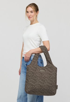 Vrouw in witte top met korte mouwen en blauwe spijkerbroek, met een grote bruine shopper met luipaardprint over haar arm tegen een effen achtergrond.