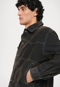 Giacca in denim nero con texture distressed, chiusura a bottoni, due tasche frontali e cuciture a contrasto lungo le cuciture.