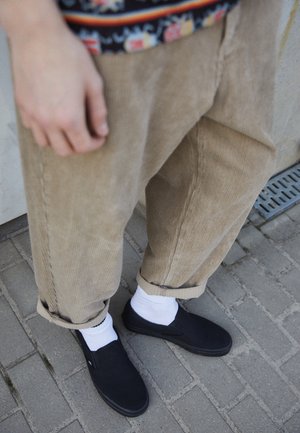 Person iført beige ribbukser, hvide sokker og sorte slip-on sko, der står på gråt fortov.