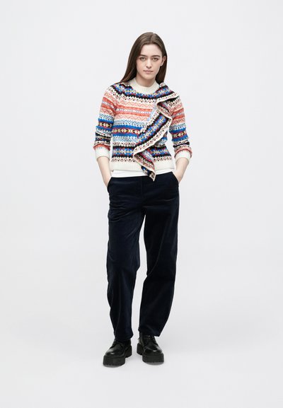 Πολύχρωμο πλεκτό πουλόβερ με μοτίβα σε κόκκινο, μπλε και εκρού, με γιακά που πέφτει χαλαρά. Συνδυάζεται με σκούρο μπλε παντελόνι κοτλέ και μαύρα παπούτσια με πλατφόρμα.