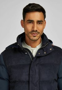 Marineblauwe pufferjack met een zachte, suede-achtige textuur. Voorzien van een ritssluiting aan de voorkant, drukknopen en een capuchon. Gestyled met een crèmekleurig shirt eronder.