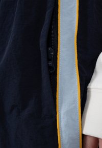 Gros plan sur la fermeture éclair partiellement ouverte d'une veste sombre, bordée d'une bande jaune et bleu clair, avec une manche blanche visible sur le côté droit.