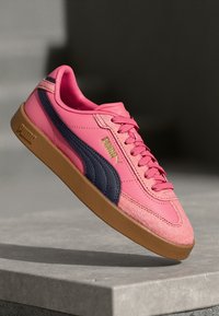 A PUMA sportcipő pink szintetikus felsőrészével, tengerészkék csíkkal, párnázott bokarésszel és barna gumitalppal rendelkezik. Texturált velúr részletek is megtalálhatók.