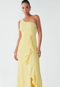 Une femme pose dans une robe jaune vif à une épaule avec un détail froncé, portant des boucles d'oreilles en or et un bracelet, devant un fond neutre.