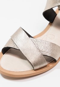 Sandale à bout ouvert avec deux larges brides métalliques croisées et une semelle beige sur fond blanc.