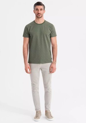 Camiseta verde de manga corta con un patrón geométrico blanco, combinada con pantalones gris claro y zapatillas beige. Cuello redondo simple y corte casual.