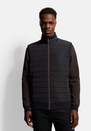 Quilted navy vest with a high collar, zippered front, and brown accents. Paired with a dark brown long-sleeve shirt. Smooth texture, athletic fit.