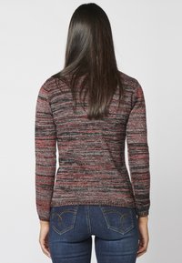 Pull en maille à rayures multicolores rouges, noires et grises, doté de manches longues et d'une coupe ajustée, associé à un jean bleu foncé.