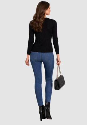 Femme aux longs cheveux bruns portant un haut noir à manches longues, un jean skinny bleu, des bottes à talons hauts noires, tenant un sac matelassé noir à bandoulières chaîne.