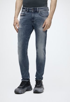 Mand iført tætsiddende falmede blå jeans og sorte chunky sneakers, stående mod en ensartet lys baggrund.