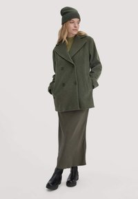 Olivgrüner Wollmantel mit doppelreihigen Knöpfen, kombiniert mit einem passenden olivgrünen Rock und schwarzen Stiefeletten. Modell trägt gestrickte Mütze.