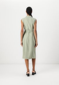 Sleeveless light green dress with a collar, belted waist, and midi length, made of textured fabric. Worn with black heeled sandals.