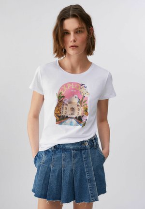 Giovane donna che indossa una maglietta bianca con il Taj Mahal, una tigre, fiori e la scritta "Explore", abbinata a una gonna di jeans blu plissettata, con le mani nelle tasche.