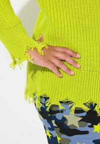 Haut en tricot vert néon vif avec des bords effilochés, associé à un pantalon camouflage bleu orné d'étoiles jaunes. La texture est décontractée et ample.