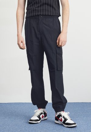 Mand iført sorte cargobukser med elastiske manchetter og hvide, sorte og røde sneakers, står på blåt tæppe mod en hvid væg.