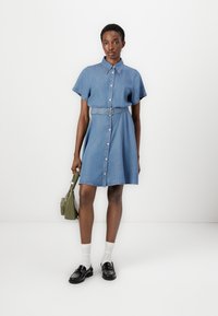Denim shirt dress in light blue, short sleeves, button front, belted waist, and above-the-knee length. Worn with black shoes and white socks.