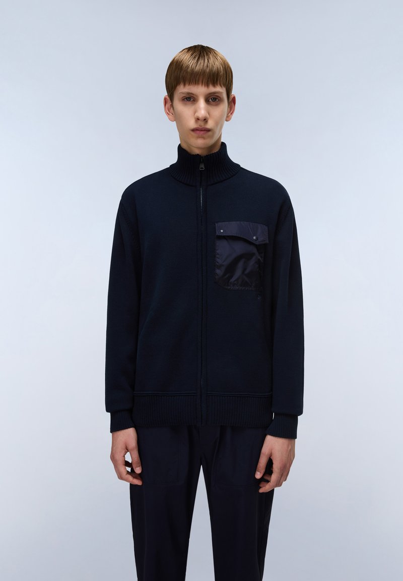 Mörkblå stickad zip-jacka med hög krage, med en framfick i en glänsande tyg, ribbade ärmslut och nederkant. Slät textur.