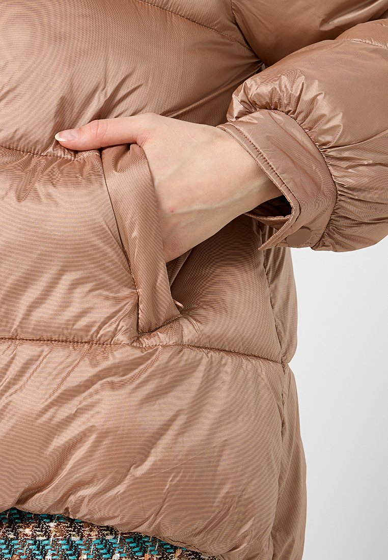 Beżowa kurtka puchowa z paskowanym wzorem, elastycznymi mankietami i boczną kieszenią. Widać rękę w kieszeni.