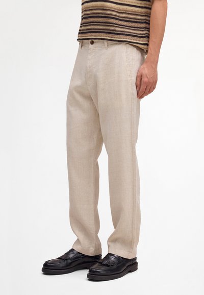 Uomo con pantaloni beige a gamba dritta, camicia a righe marroni e mocassini di pelle nera, in piedi contro uno sfondo bianco semplice.