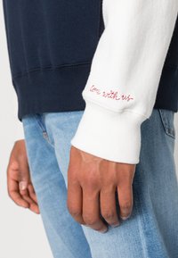 Marineblaues und weißes Sweatshirt mit gerippten Bündchen; mit rotem, gesticktem Text "Komm mit uns" auf dem weißen Ärmel. Weicher Stoff und lockere Passform.