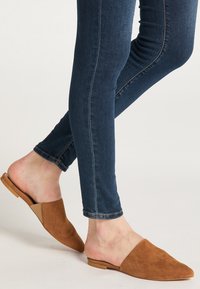 Braune Wildleder-Mules zum Hineinschlüpfen mit spitzem Zehendesign. Getragen mit engen Jeans aus dunklem Denim, die eine glatte Textur und eine schlichte Silhouette zeigen.