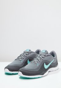 Zapatos deportivos grises con parte superior de malla, acentos turquesa, suela blanca y un acabado texturizado en los lados y la puntera. Logo de Nike visible.