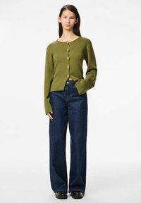 Oliņa zaļā džemperis ar pogām, gariem piedurknēm un nedaudz īsu dizainu, apvienots ar brīviem tumši zilajiem džinsiem un melniem apaviem.