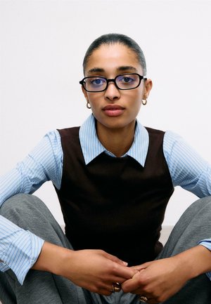 Mladá žena s brýlemi, oblečená v černé vestě přes modrou pruhovanou košili a šedé kalhoty, sedící s nohama zkříženýma na jednobarevném bílém pozadí.