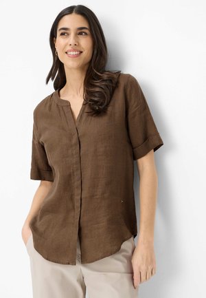 Junge Frau mit langem dunklem Haar, die eine braune kurzärmelige Bluse und hellbeige Hosen trägt, lächelt und vor einem weißen Hintergrund steht.