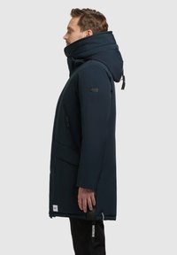 Parka isolata blu navy con colletto alto, cappuccio rimovibile e tasche laterali con zip. Presenta un patch con logo sulla manica e cordini regolabili.