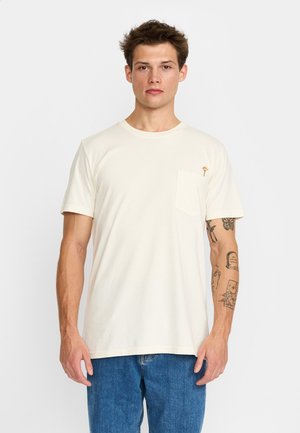 Young man with short brown hair wearing a cream t-shirt with a small flower in the chest pocket and blue jeans, standing against white background.