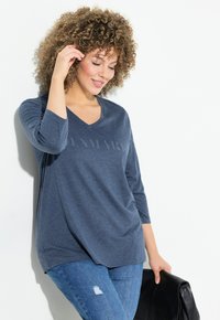 Blaues V-Auschnitt Langarm-T-Shirt mit einem dezenten Logo auf der Vorderseite, kombiniert mit hellblauen, verwaschenen Jeans und einer schwarzen Handtasche.