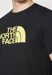 Černé bavlněné tričko s velkým žlutým logem "The North Face" napříč hrudníkem, s kulatým výstřihem a krátkými rukávy.