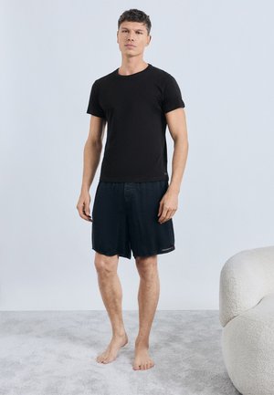 Zwart T-shirt met korte mouwen, gecombineerd met donkerblauwe denim shorts. Model staat op blote voeten op een lichtgrijs tapijt, minimalistische achtergrond.