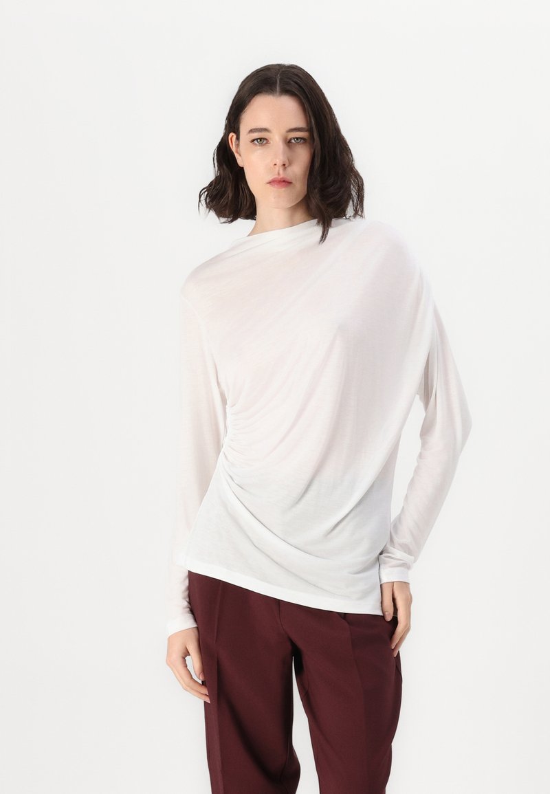 White long-sleeve top with a draped design, featuring a round neckline and gathered detail on the side, paired with dark maroon trousers.