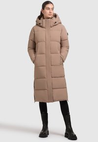 Beige puffatakki hupulla, jossa on pitkä, suora muotoilu ja vaakasuora tikkauskuvio. Vetoketjulliset sivutaskut ja minimaalinen yksityiskohtaisuus.