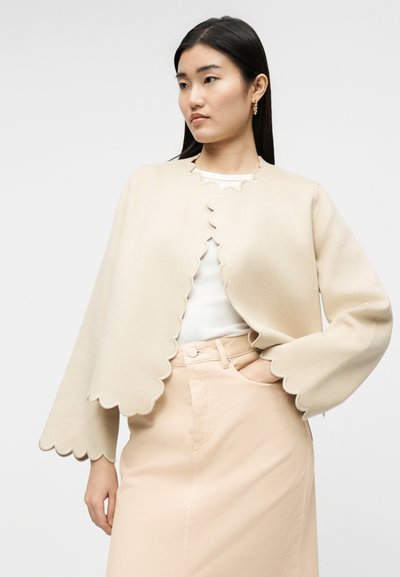 Γυναίκα με μακριά μαύρα μαλλιά που φοράει μπέζ καπέλο-πάνω ζακέτα με κυματιστό τελείωμα, λευκό τοπ και μπέζ ψηλόμεση φούστα, στέκεται με το ένα χέρι στην τσέπη.