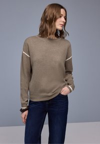 Femme en pull beige avec des rayures blanches sur les manches et un jean bleu foncé, debout avec une main dans la poche devant un fond gris.