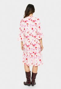 La robe fleurie présente un tissu rose clair avec des fleurs rouges et violettes, des manches longues, une jupe à volants, et des bottes brunes jusqu'aux genoux.