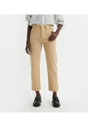 Personne portant un jean beige taille haute à jambe droite, une chemise blanche à rayures avec les manches retroussées, et des mocassins noirs sur un fond blanc.