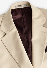 Blazer beige con un exterior texturizado y un forro de patrón marrón. Cuenta con solapas afiladas y un pequeño bolsillo decorativo en la parte delantera.