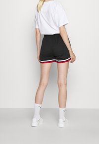 Nainen seisoo selkä menosuuntaan, päällään mustat urheilushortit, joissa on punaiset ja valkoiset raidat, valkoinen t-paita, valkoiset sukat ja valkoiset lenkkitossut.